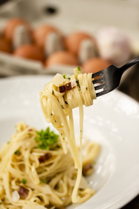 Spaghetti Carbonara - Spaghettinudeln aufgewickelt auf einer Gabel, Käseraspeln und Speckwürfel sind zu erkennen. Im Hintergrund befindet sich ein weißes Teller mit einer Portion Spaghetti Carbonara. (Foto: Tobias Schneider-Lenz - Nicht zur freien Verwendung)